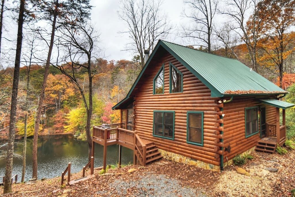 A log cabin with a green roof, a deck, and a pond in the background.