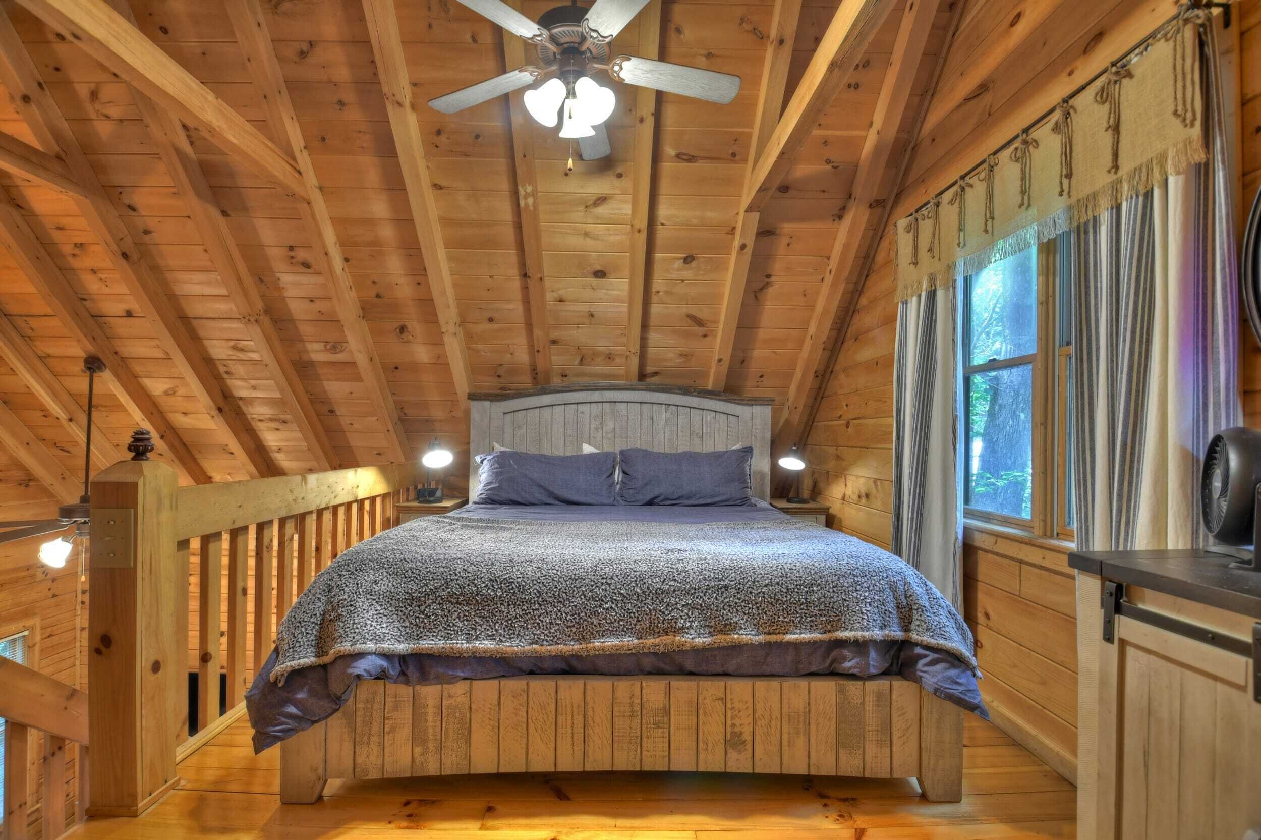 Un dormitorio de cabaña de madera con una cama, un ventilador de techo y una ventana con cortinas.