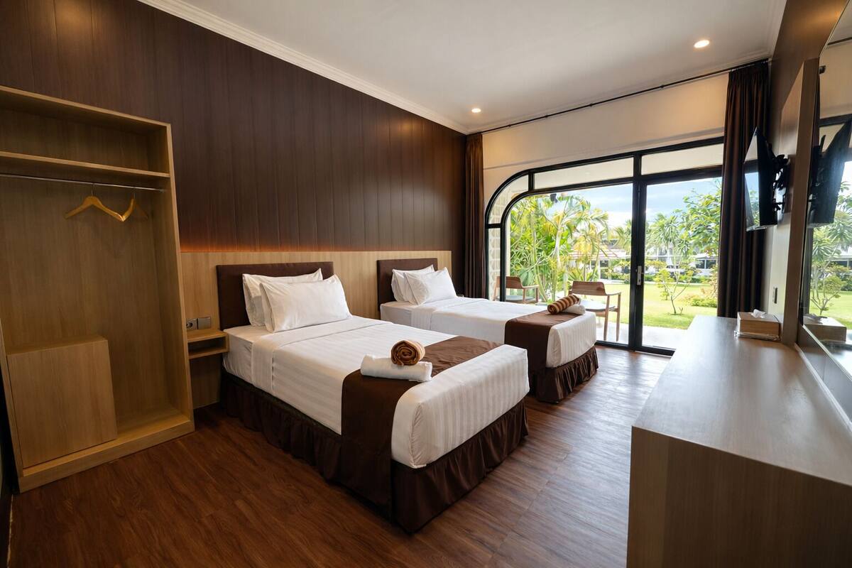 Habitación de hotel con dos camas, un armario de madera, un escritorio y un amplio ventanal con vistas a zonas verdes.