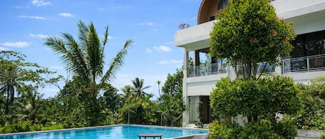 Un área de piscina con sillones de descanso, rodeada de abundante vegetación y un edificio moderno con ventanas arqueadas.