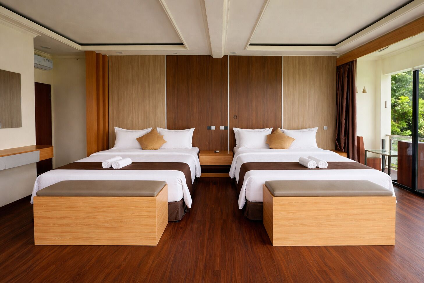 A modern hotel room with two beds, wooden flooring, and large windows offering a view of greenery.