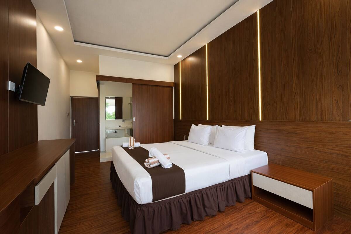 Habitación de hotel moderna con una cama grande, paneles de madera, un televisor de pantalla plana y un baño visible a través de una puerta abierta.