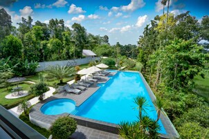 Una piscina rectangular con una sección curva, rodeada de sillas de descanso y sombrillas, ubicada en un exuberante jardín con árboles altos y una pared blanca al fondo.