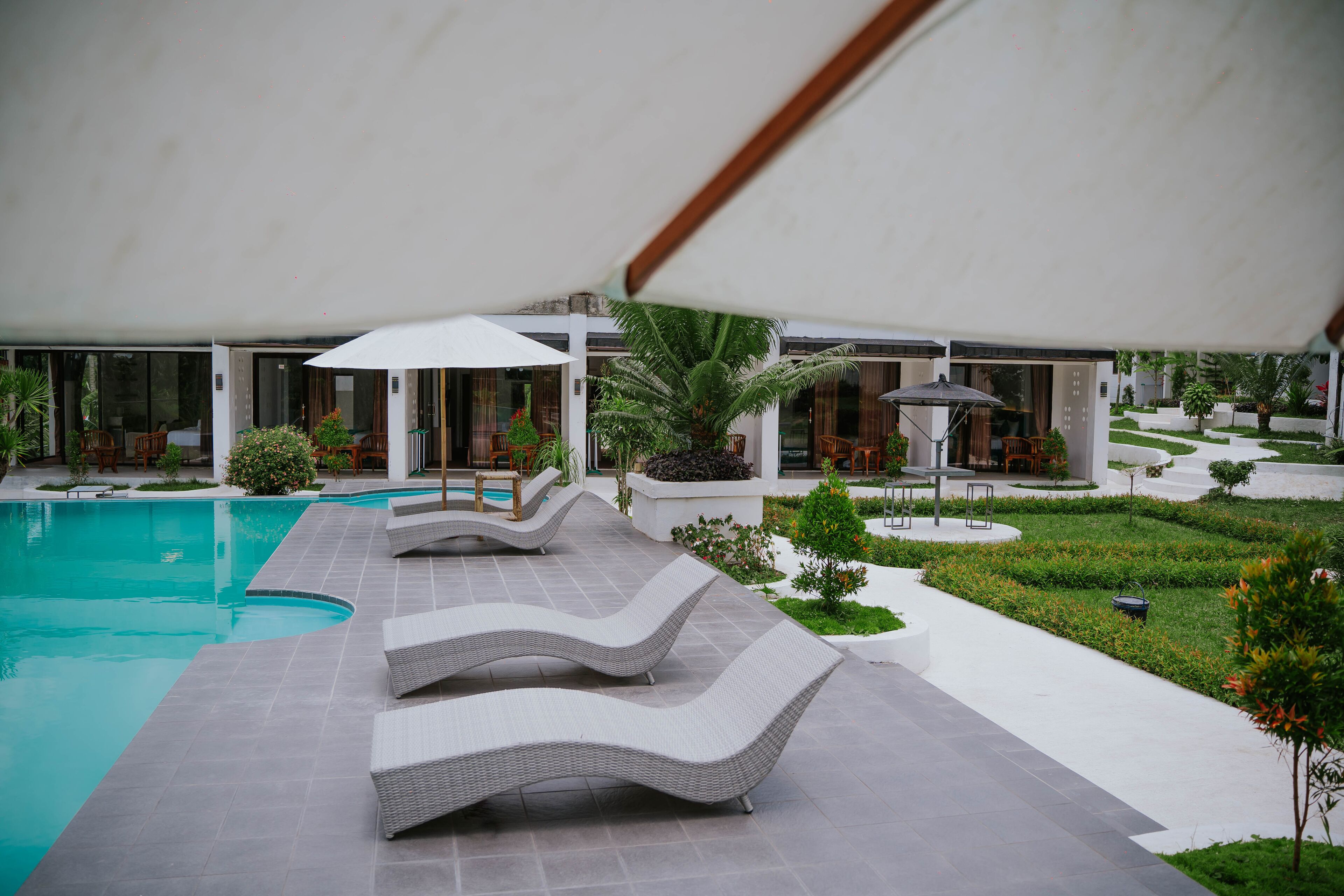 Een modern buitenzwembad met ligbedden en parasols, omringd door verzorgde beplanting en een gebouw met grote ramen.