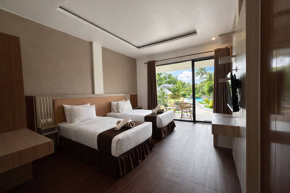 Habitación de hotel con dos camas, televisión, escritorio y vistas a una piscina y vegetación.