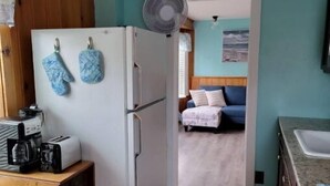 Cozinha compacta com geladeira branca, mesa de madeira com cafeteira e um ventilador. Uma cama com cabeceira azul é visível ao fundo.