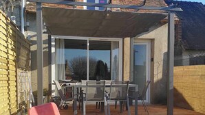 A covered patio area with a table and chairs, a red and blue lounge chair, and a view of the house.