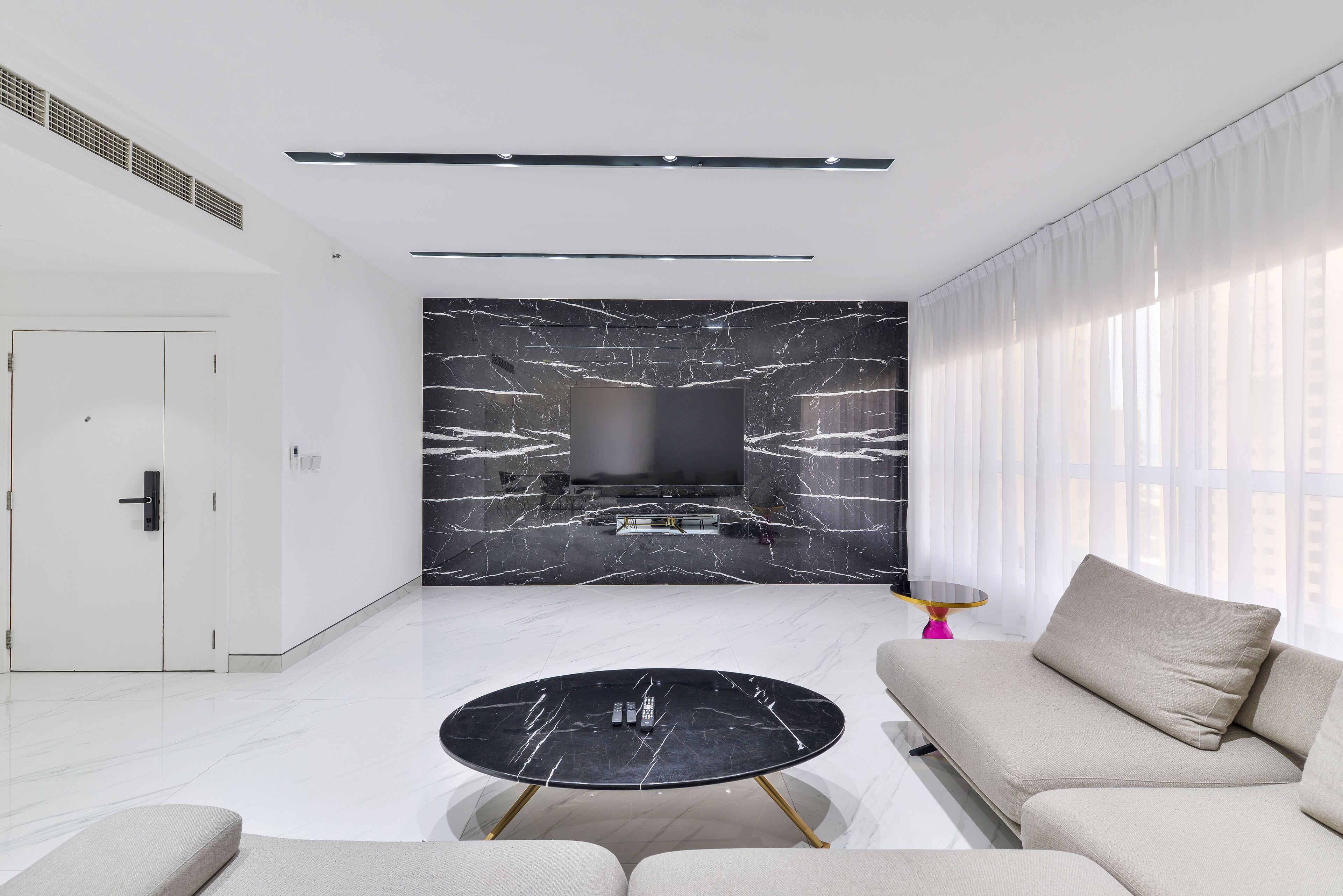 A modern living room with a flat-screen TV mounted on a marble wall, a round black coffee table with gold legs, and a beige sofa.