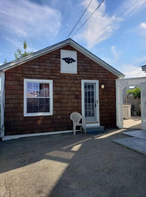 Un petit bâtiment en bois avec une porte et des fenêtres blanches, une chaise blanche à l’extérieur et un logo sur le mur.