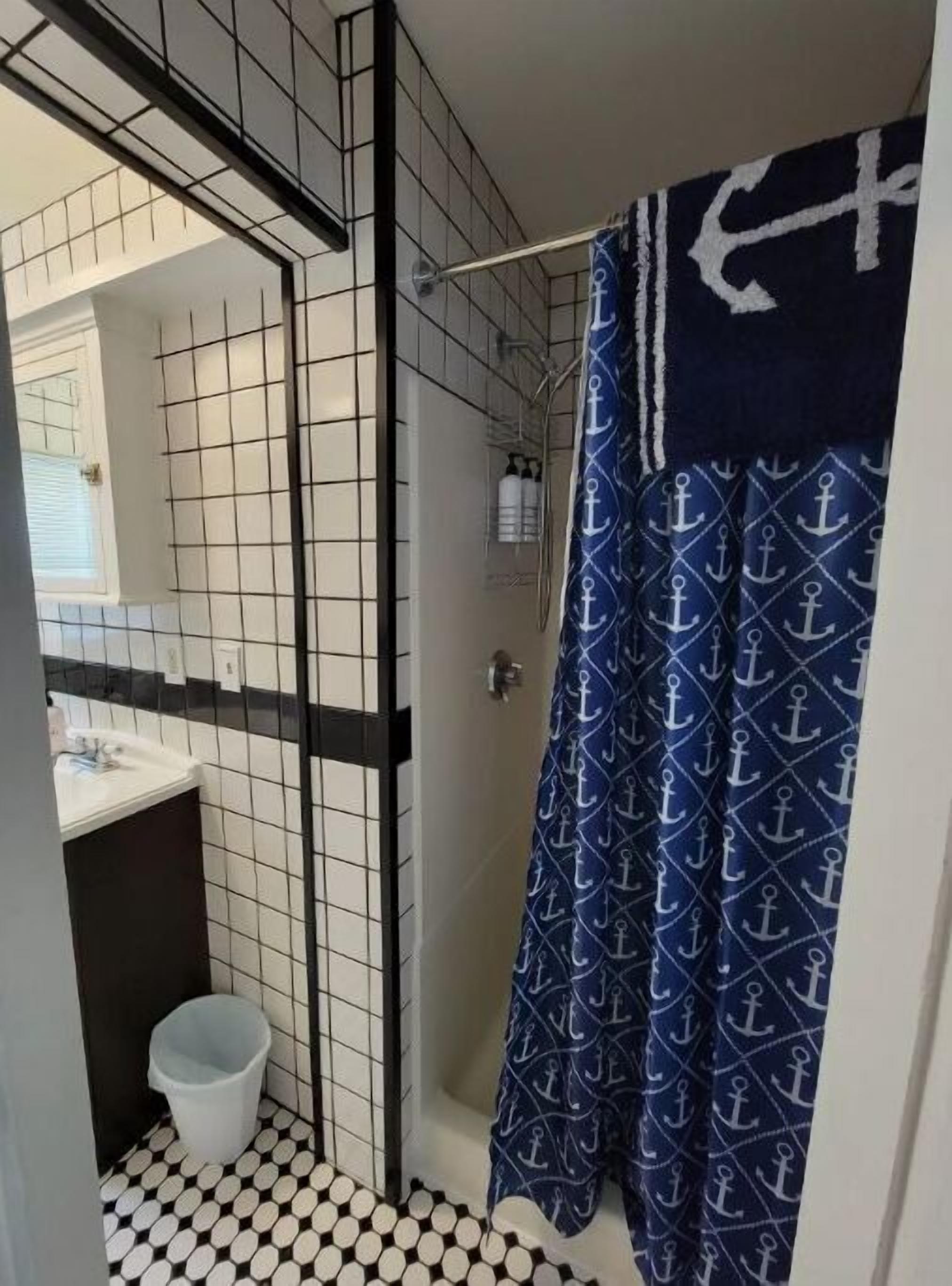 Un baño con piso de baldosas, una ducha con cortina azul con motivos de anclas, un lavamanos blanco y un espejo.