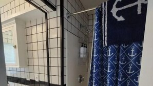 Een badkamer met betegelde vloer, een douche met blauw gordijn met ankerprint, een witte wastafel en een spiegel.