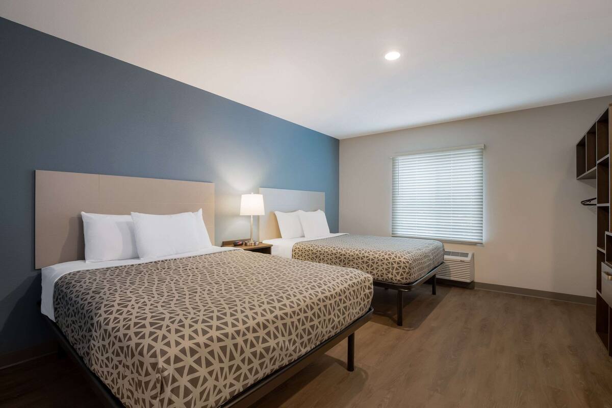 A hotel room with two beds, a nightstand with a lamp, a window with blinds, and a built-in bookshelf.