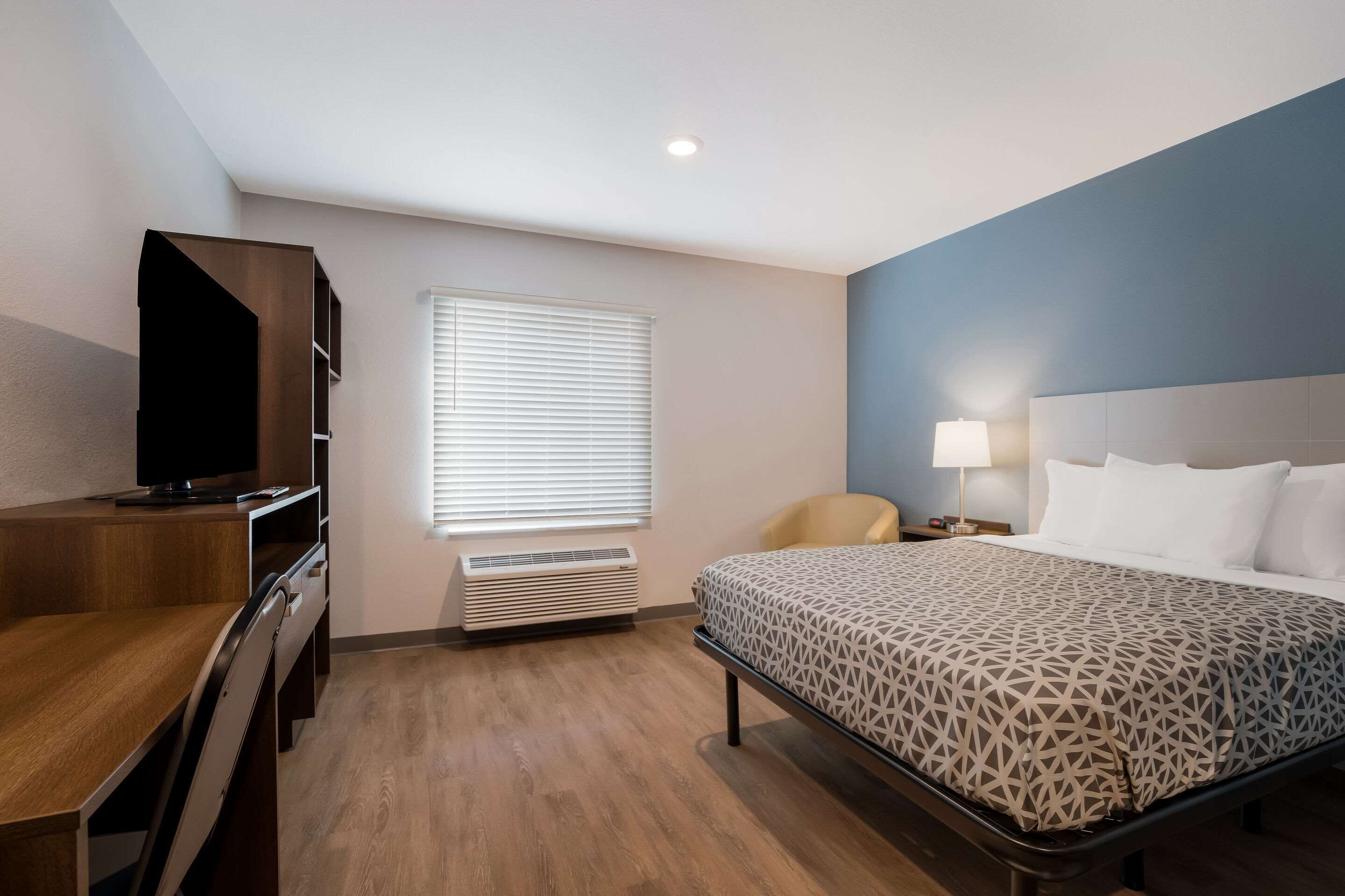 Quarto de hotel com cama, escrivaninha, televisão, cadeira e janela com persianas.