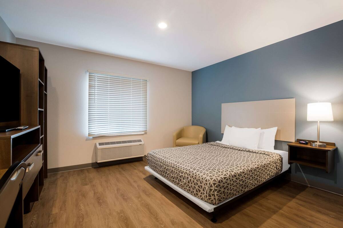 A hotel room with a bed, a chair, a TV, a heater, and a nightstand with a lamp.