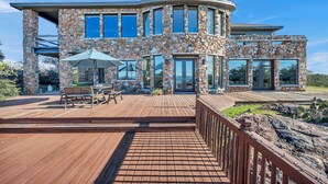 A stone-clad house with a wooden deck, outdoor seating, and a large window overlooking the landscape.