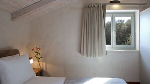 Een slaapkamer met een bed, nachtkastje, lamp, raam met gordijnen en uitzicht op bomen.