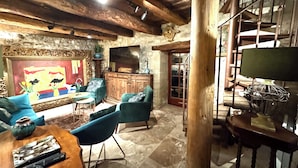 Un interior rústico con vigas de madera a la vista, pared de piedra, televisor grande y una mezcla de muebles modernos y tradicionales.