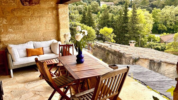 Un patio al aire libre con pared de piedra, un sofá blanco, sillas de madera y una mesa con un jarrón de flores.