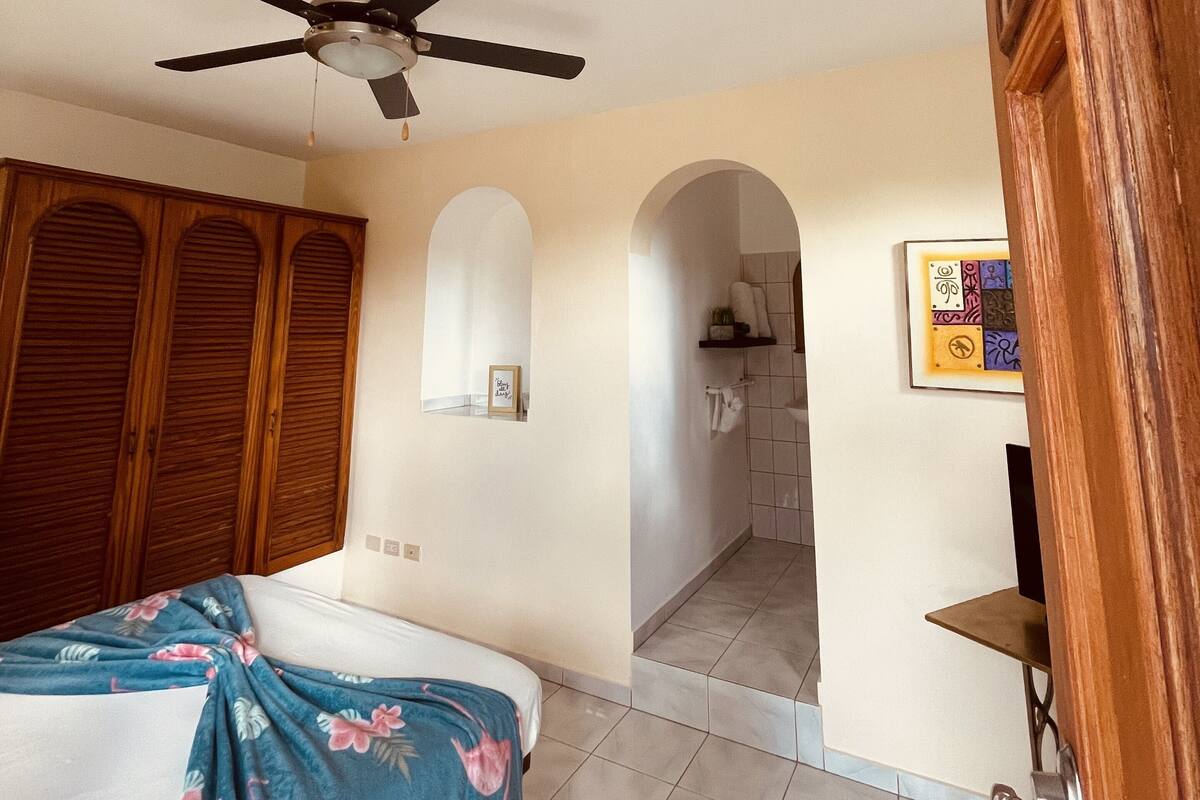 A bedroom with a bed, a ceiling fan, wooden wardrobe, and a bathroom visible through an archway.
