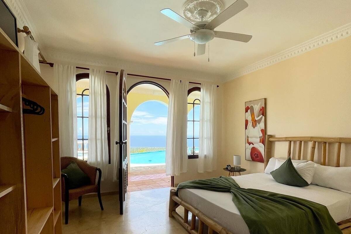 A bedroom with a bed, a chair, a wardrobe, and a view of the ocean.