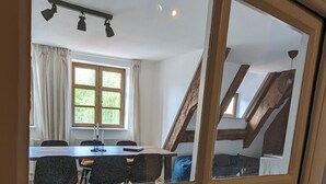 Zona de menjador amb taula i cadires de fusta, una finestra amb cortines i una escala que puja a un nivell superior.