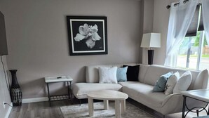 A living room with a sofa, ottoman, and a coffee table. There is a framed picture on the wall and a lamp beside the sofa.