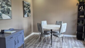 A dining area with a white table, four chairs, a grey cabinet, and a black shelving unit with plants.