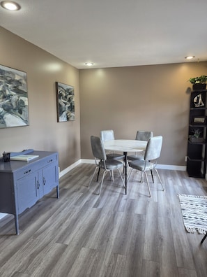 A dining area with a white table, four chairs, a grey cabinet, and a black shelving unit with plants.