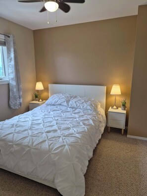 A bedroom with a bed, two bedside lamps, a ceiling fan, and a window with curtains. - Beach Retreat with Hot Tub | 1 Block to Beach | Free Parking (Sylvan Lake)