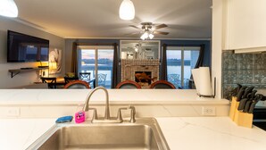 A modern kitchen with a stainless steel sink, a ceiling fan, and a view of a fireplace and balcony. - Waterfront Condo: Stunning Sunsets, Fishing, Pool, Private Patio, Near Beach  (Corpus Christi)