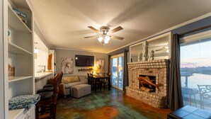 A living room with a fireplace, a ceiling fan, and a large window with a view. - Waterfront Condo: Stunning Sunsets, Fishing, Pool, Private Patio, Near Beach  (Corpus Christi)