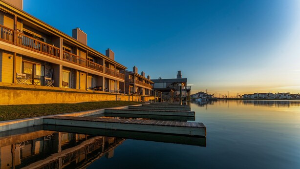 A waterfront hotel with a dock extending into calm water, featuring multiple balconies and a serene sunset view. - Waterfront Condo: Stunning Sunsets, Fishing, Pool, Private Patio, Near Beach  (Corpus Christi)