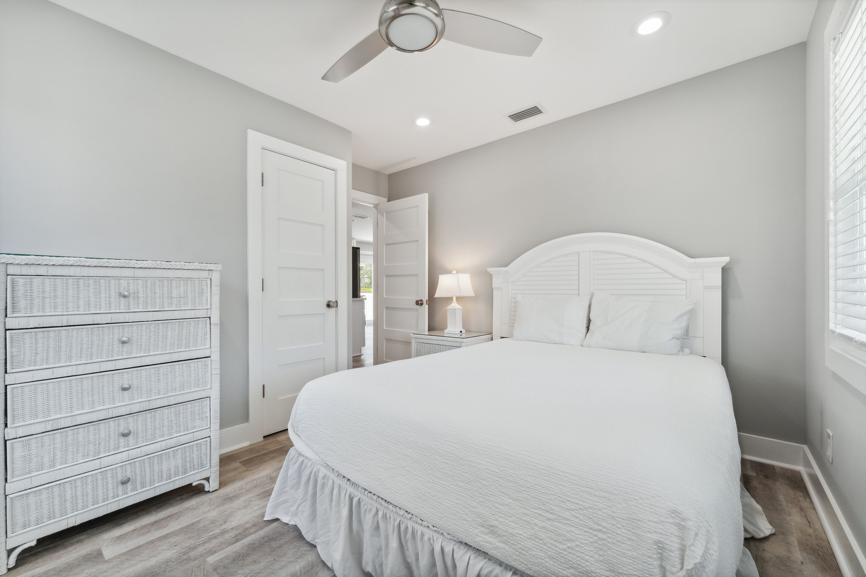 A bedroom with a large bed, a dresser, a ceiling fan, and a nightstand with a lamp.