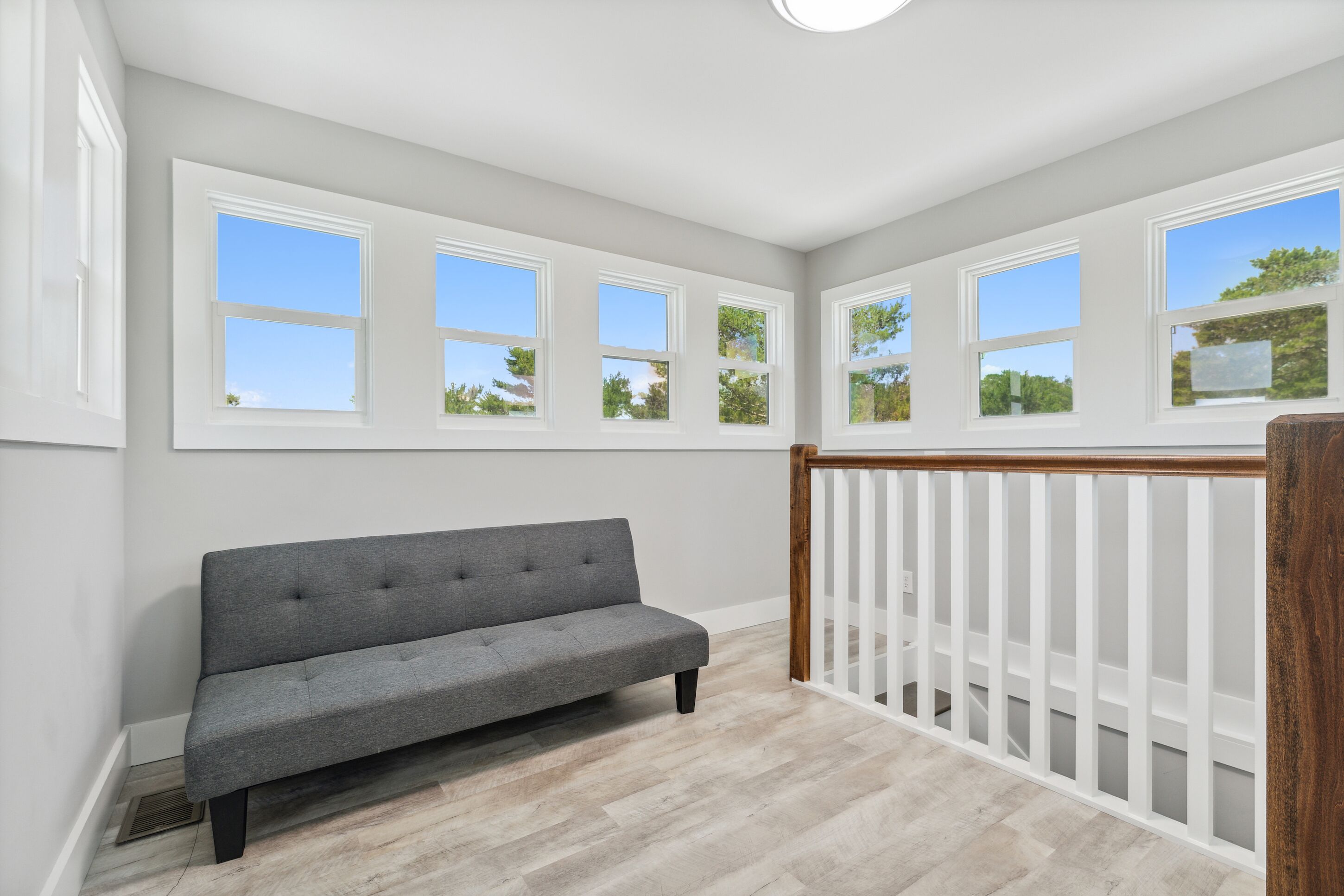 A room with a grey sofa, white walls, and multiple windows offering views of trees and sky.