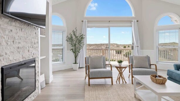 A spacious living room with a fireplace, large windows, and a view of the landscape. - Relaxing Oceanfront Retreat | Pool, Hot Tub & Beach - Sleeps up to 14! (Rodanthe)