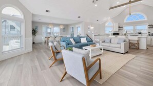 A spacious living area with a large sofa, a coffee table, and a dining table with chairs. There is a kitchen area with modern appliances and a large window. - Relaxing Oceanfront Retreat | Pool, Hot Tub & Beach - Sleeps up to 14! (Rodanthe)