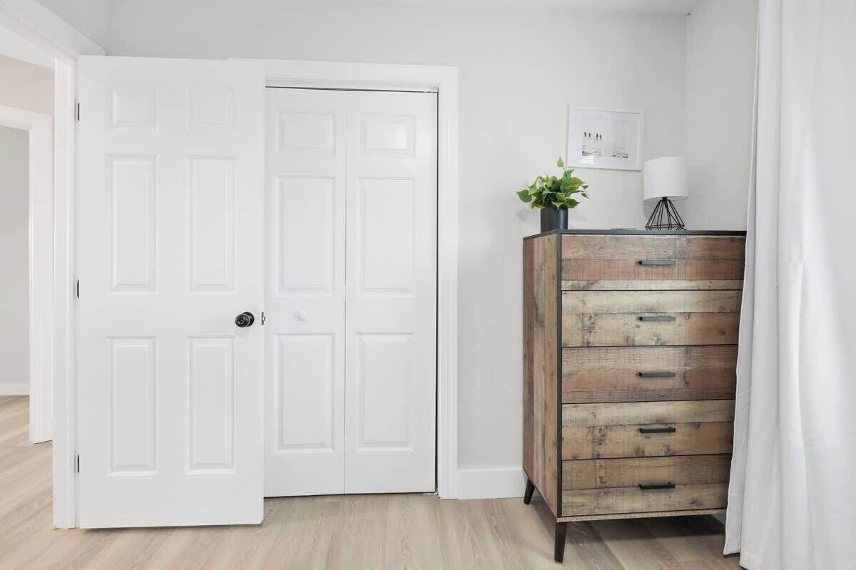 Een kamer met een witte deur, een houten dressoir met lades, een klein plantje erbovenop en een lijst met een afbeelding aan de muur.