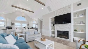 A spacious living room with a large sofa, a coffee table, and a fireplace. There is a kitchen area with white cabinets and a flat-screen TV mounted on the wall. - Relaxing Oceanfront Retreat | Pool, Hot Tub & Beach - Sleeps up to 14! (Rodanthe)