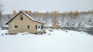 Una casa de dos pisos con porche delantero y una pequeña terraza, rodeada de árboles cubiertos de nieve y un lago.