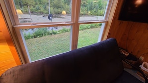 Una habitación con un ventanal grande que da a una zona boscosa, suelo de madera y un sofá de color oscuro.