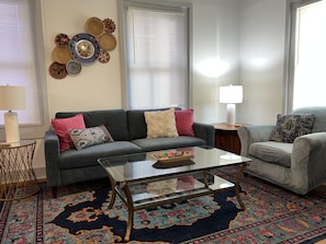 Un salon avec un canapé gris, une table basse en verre, un tapis à motifs et un mur décoré d’assiettes.