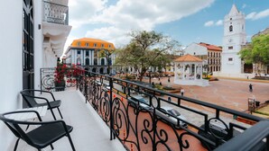 A balcony with black metal chairs, a white building with an orange roof, and a plaza with a white clock tower. - Lefevre 1C (Panama City)