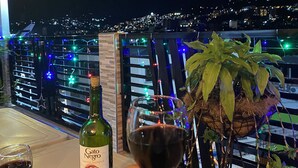 Une main tenant un verre à vin, avec une bouteille de vin et une plante sur un balcon donnant sur une ville la nuit.