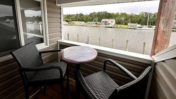 Een balkon met rieten stoelen en een tafeltje, met uitzicht op een meer met boten en huizen.