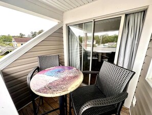 Un balcon avec une table ronde et deux chaises en osier, une porte-fenêtre coulissante et une vue sur un quartier résidentiel.