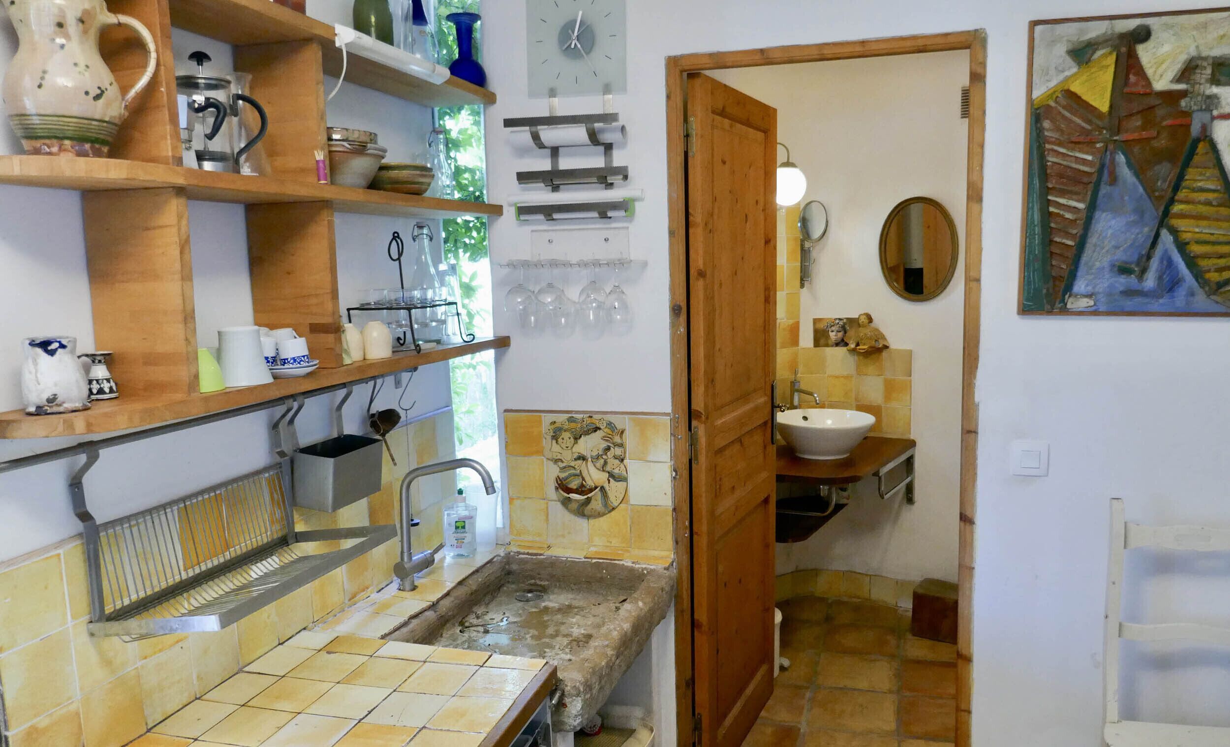 A compact kitchen with a sink, shelves, and a wooden door leading to a bathroom with a mirror and tiled walls.