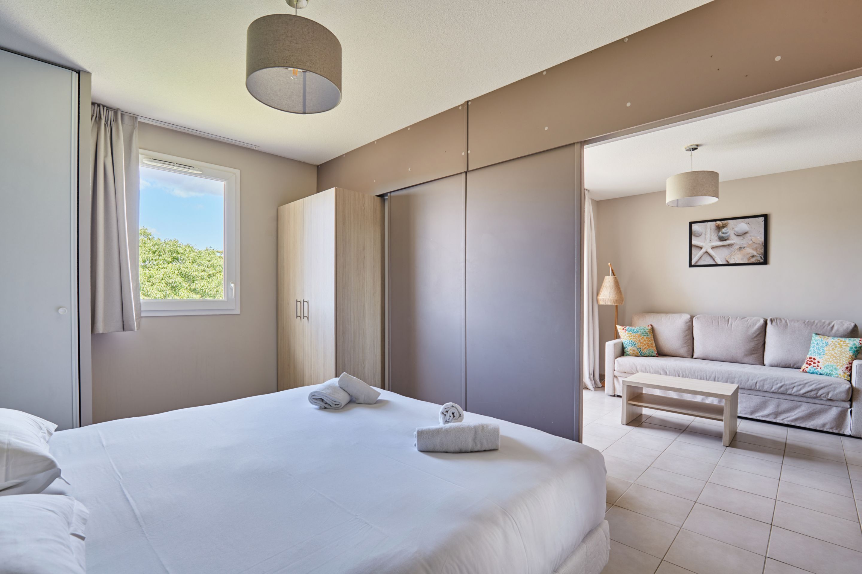 Une chambre d’hôtel moderne avec un grand lit, un canapé et une table à manger. Une fenêtre laisse entrer la lumière naturelle et offre une vue sur la verdure environnante.