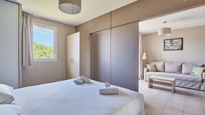 A modern hotel room with a large bed, a sofa, and a dining table. There is a window providing natural light and a view of greenery outside. - Residence Odalys Les Oceanides (La Londe-les-Maures)