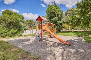 Un parque infantil con tobogán y pared de escalar, rodeado de árboles y superficie de grava.