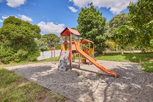 Un parque infantil con tobogán y pared de escalar, rodeado de árboles y superficie de grava.
